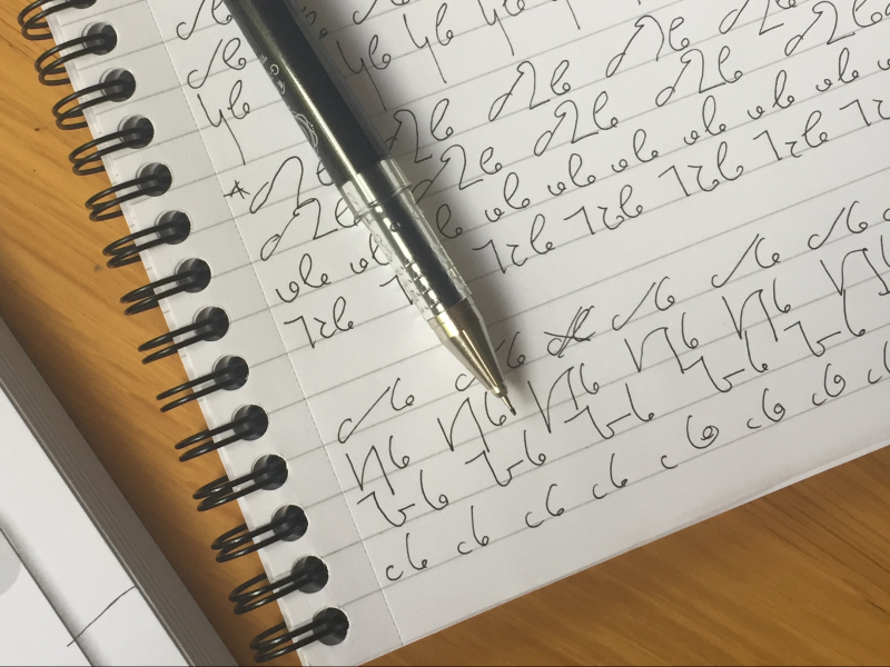 A notepad is on a wooden table and has a pen resting on top of it. On the pages there are lots of shorthand notes. On the left hand side of the photo is the corner of the shorthand exercise book.